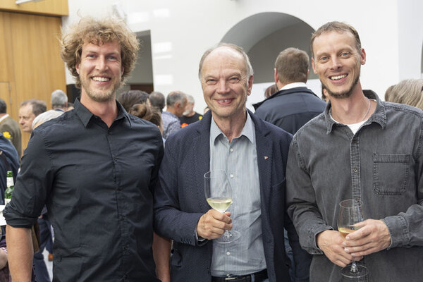 Drei Männer. Zwei halten ein Glas Wein in der Hand