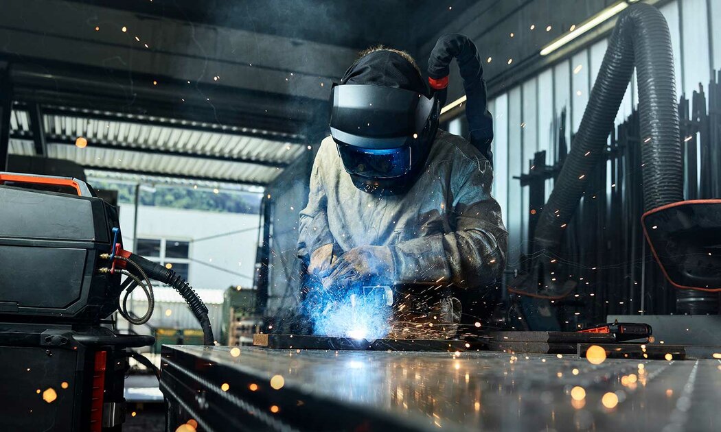 Ein Mann arbeitet in einer Fabrik und schweißt Metallteile, während Funken sprühen und Maschinen im Hintergrund stehen.