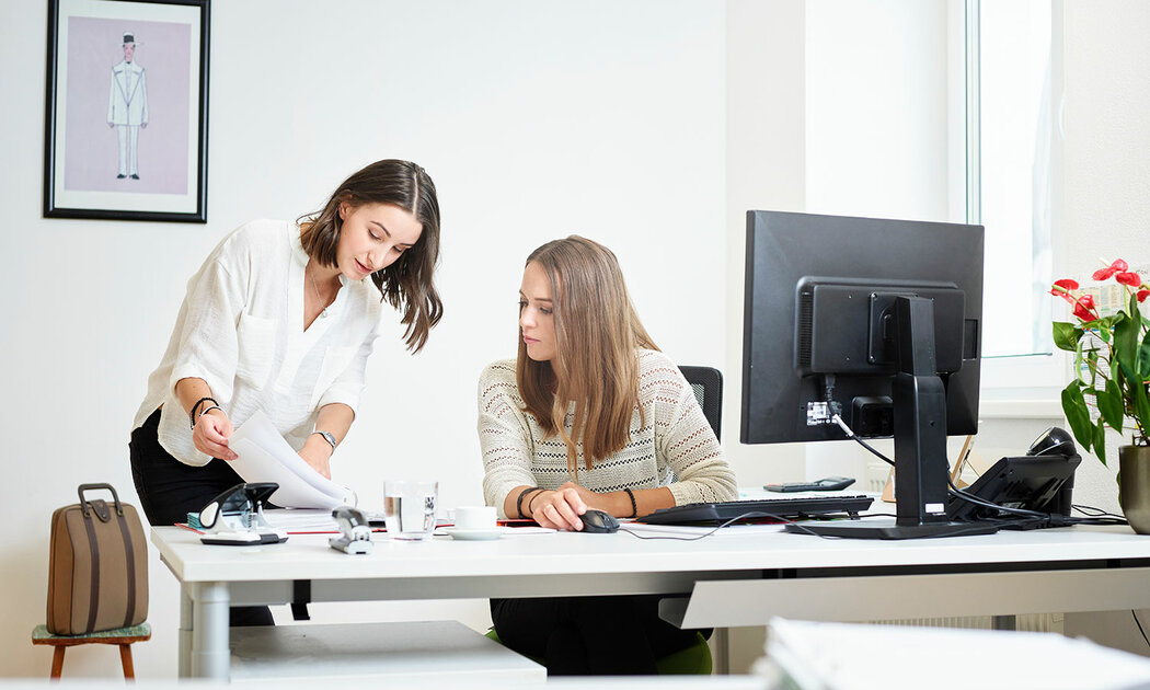 Zwei Frauen am Schreibtisch in einem Büro, die zusammen an ihren Aufgaben arbeiten und sich austauschen.